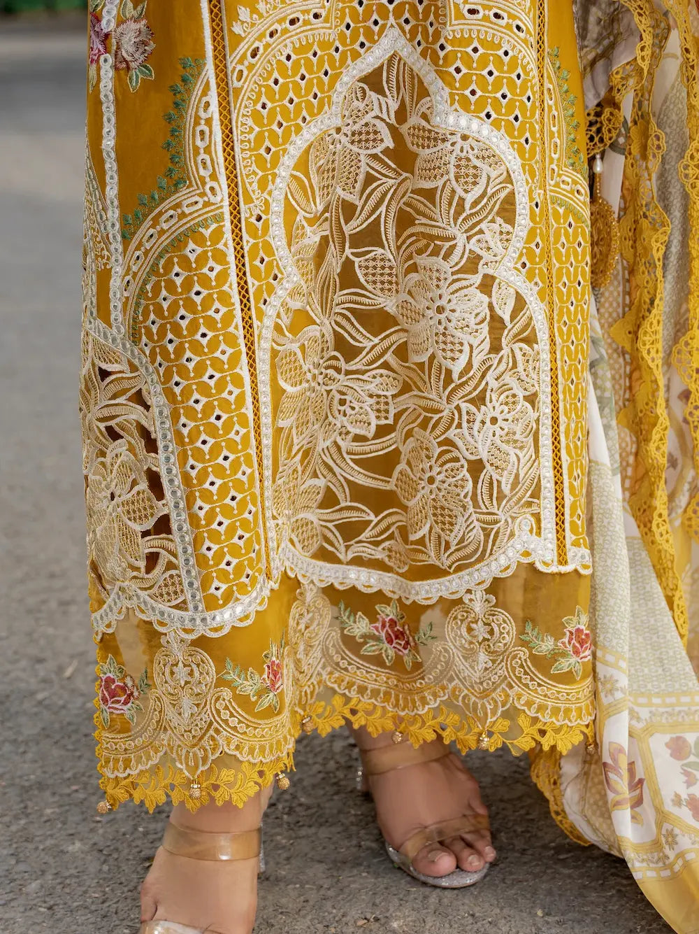 Close-up of a mustard yellow embroidered skirt with intricate white floral lace cutwork and scalloped borders, worn with clear high-heel sandals.