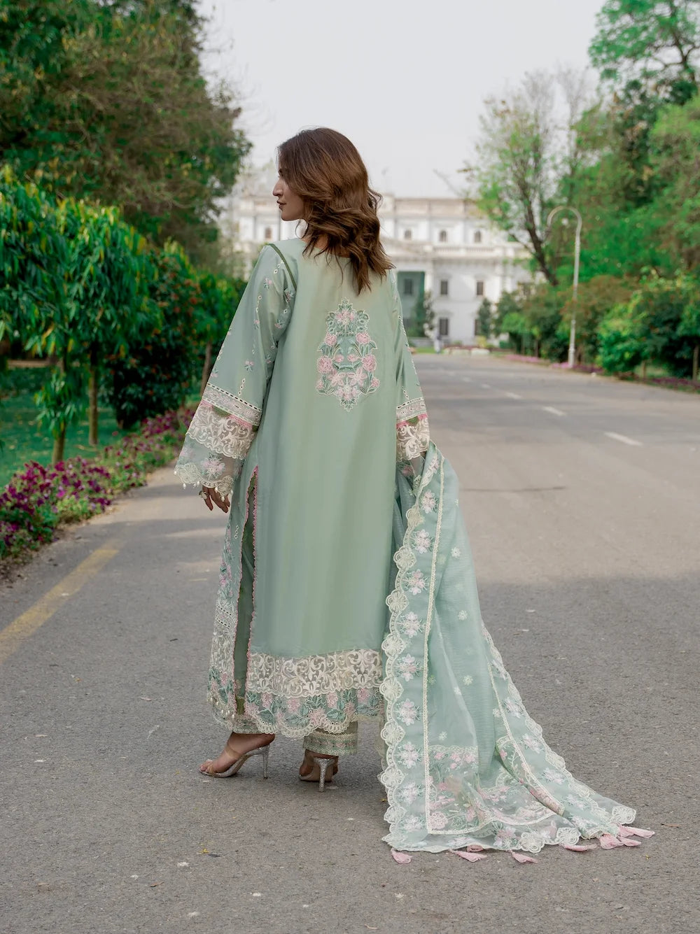 Woman in a light green embroidered traditional outfit walking on a road with greenery around.
