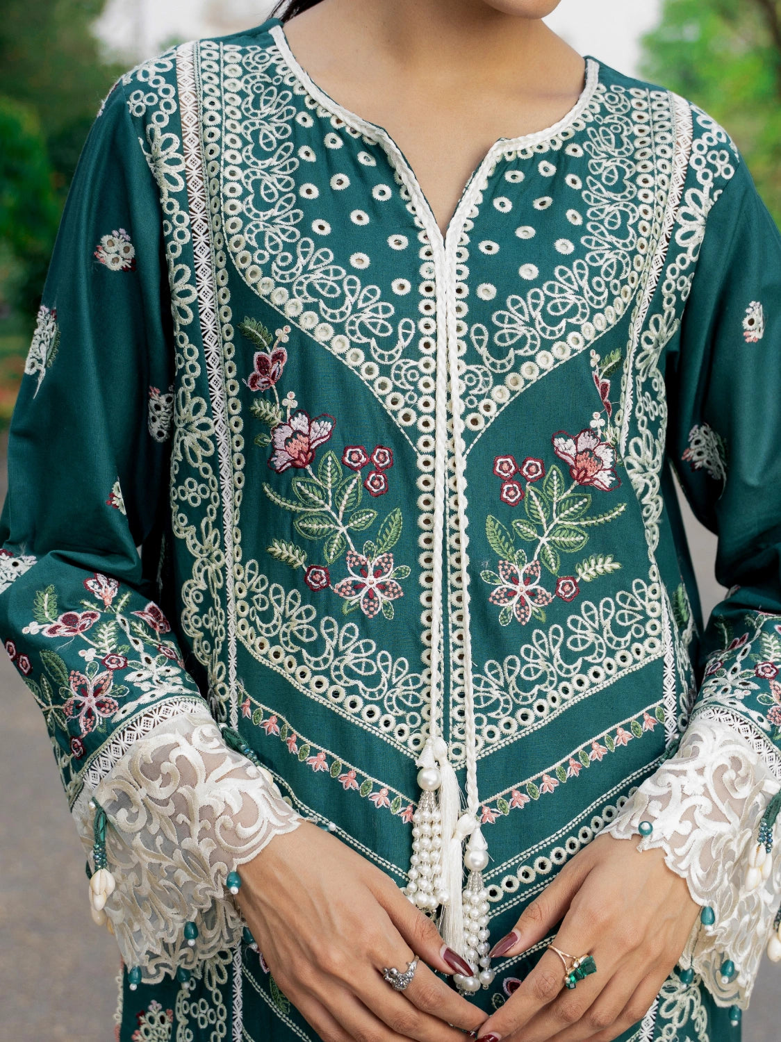A close-up of a dark teal green embroidered tunic with intricate white lace-like embroidery, floral motifs, and tassel details, worn by a model whose hands and lower face are visible.