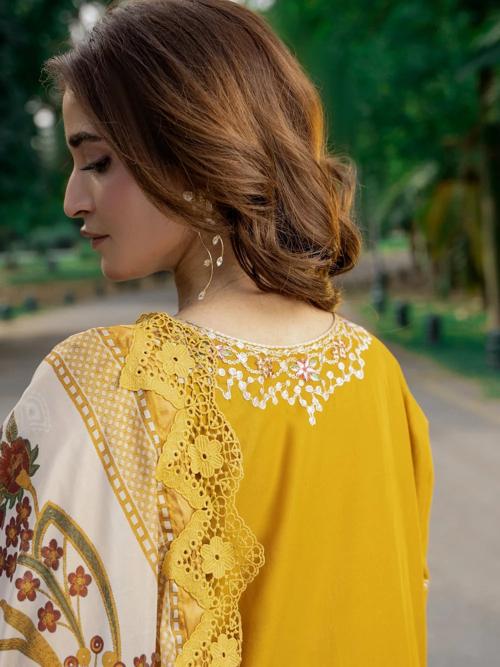 A woman wearing a yellow traditional outfit with intricate white and gold embroidery around the neckline and a matching patterned dupatta, photographed outdoors on a blurred path with greenery in the background.