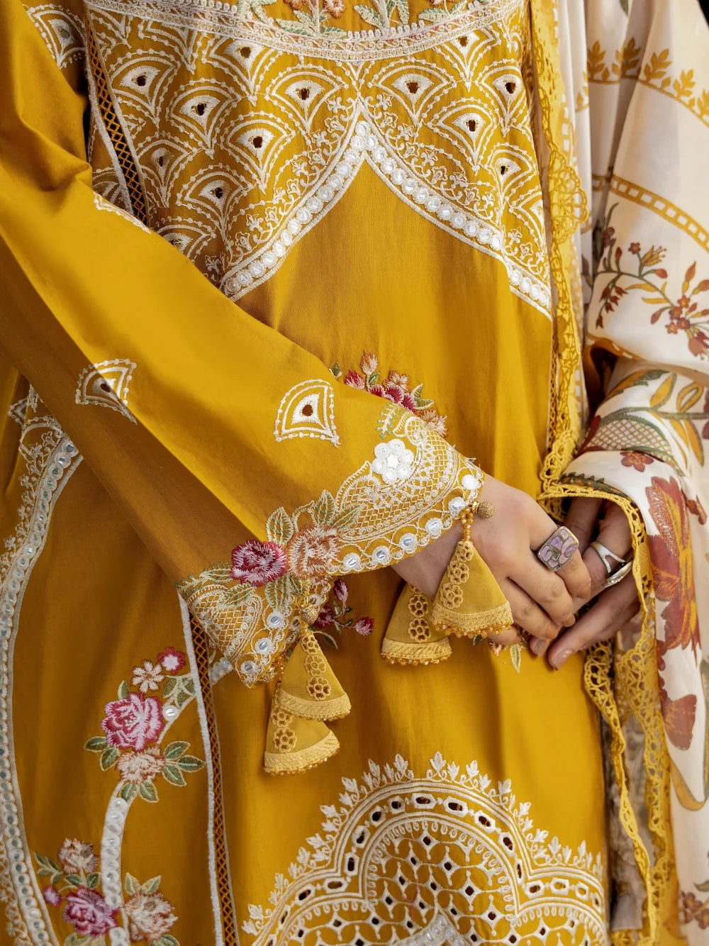 A close-up of a mustard yellow traditional embroidered outfit with intricate white and gold geometric patterns, floral embroidery, and tassel details, worn by a model whose hands are clasped in front.