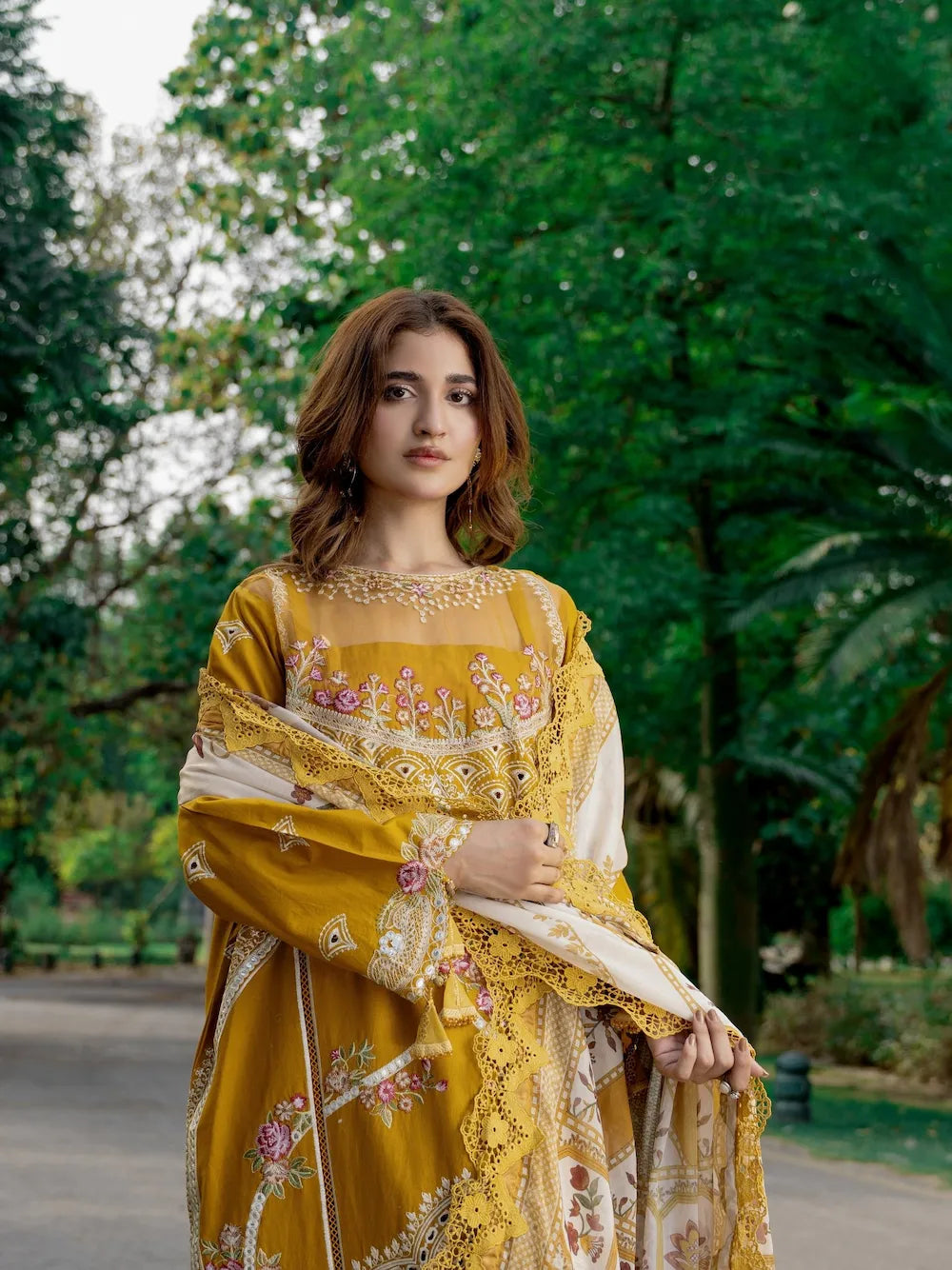 A woman wearing a traditional embroidered yellow and white salwar kameez suit with a matching dupatta, standing outdoors in a park with green trees in the background.