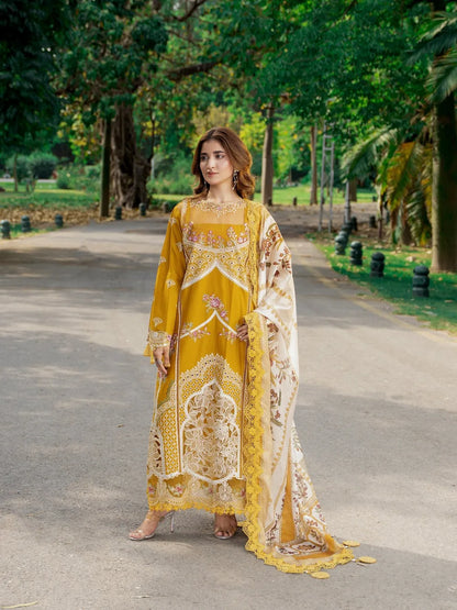 A woman stands on a paved path in a park wearing a traditional embroidered outfit. The outfit is a mustard yellow long kurta with intricate cream and gold embroidery, paired with a matching cream dupatta featuring gold floral patterns.