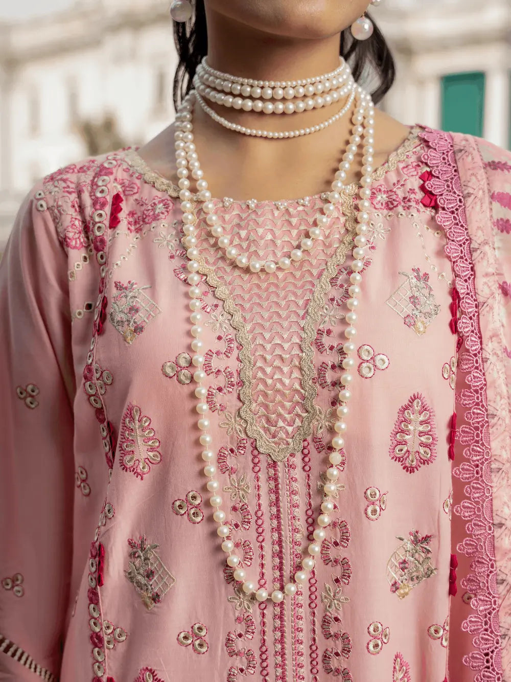 A close-up of a person wearing a light pink embroidered traditional outfit with intricate floral and geometric patterns, layered with multiple strands of long pearl necklaces.
