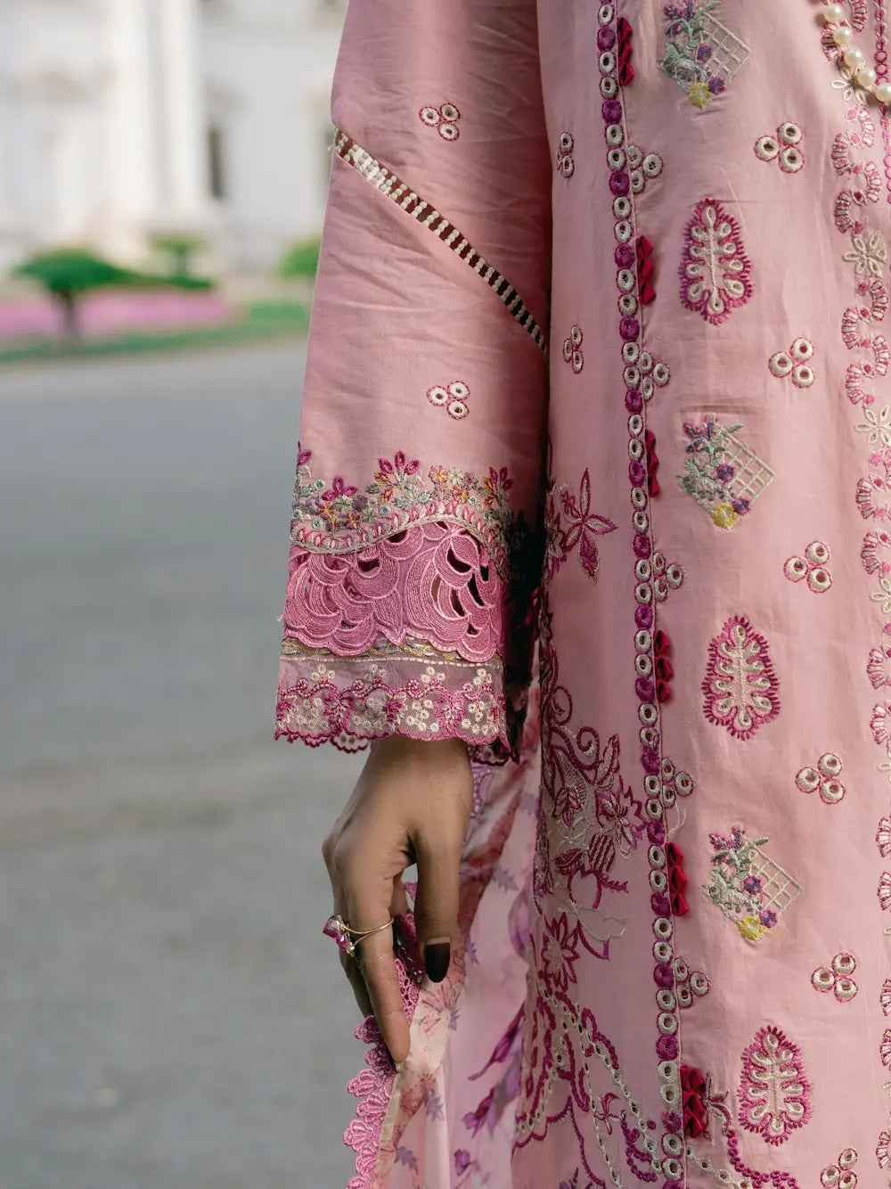 A close-up of a pink embroidered traditional garment with ornate floral and geometric embroidery, featuring a decorative scalloped sleeve cuff and a hand holding the fabric.