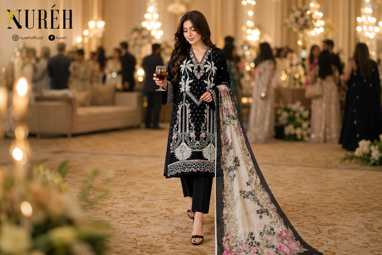 A woman walks down a grand indoor hallway wearing a black embellished kurta with silver embroidery, black trousers, and a long beige printed dupatta with floral patterns. The Nuréh brand logo is visible in the top left corner.